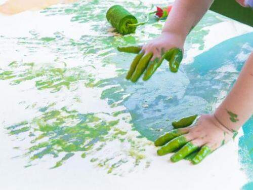 criança pintando painel verde com tinta nas mãos 