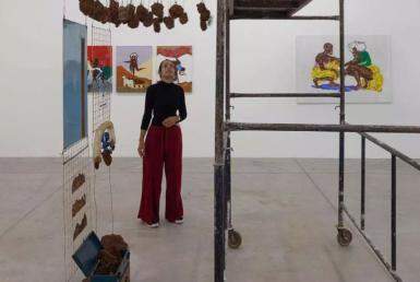 A imagem mostra uma mulher usando calça vermelha, blusa preta em uma galeria de arte. Vários quadros estão pendurados nas paredes