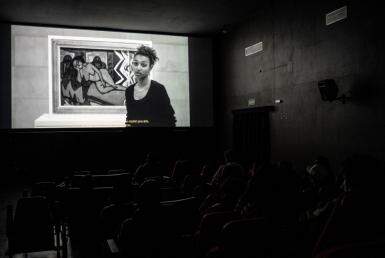 A imagem mostra pessoas em uma sala de cinema. Na tela aparece uma mulher negra. 