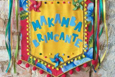 A imagem mostra o estandarte do Makamba, em tecido de xita, com um fundo amarelo e escrito em azul Makamba Brincante