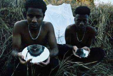 Dois jovens negros seguram, cada um, um espelho enquanto olham para câmera