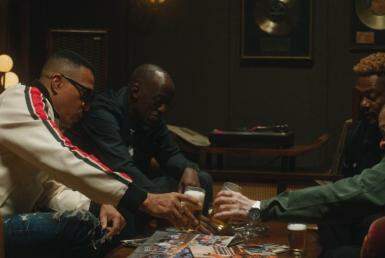 Quatro homens negros brindam com copos de cerveja em uma mesa baixa