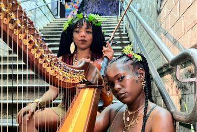 Duas mulheres negras, segurando uma harpa, olham para câmera enquanto estão sentadas em uma escada. Elas usam algumas plantas no cabelo, como adereço 