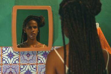 Mulher negra olhado para um espelho fixado na parede. Ela usa tranças. Na imagem só é possível ver o rosto dela pelo reflexo do espelho