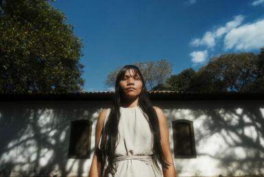 mulher indígena, olhando para câmera. Ela está em pé, com os cabelos longos e soltos, franja e vestido cinza. Ao fundo há uma casa branca, um pedaço do céu azul e árvores na lateral