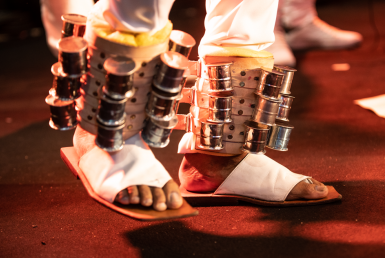 A imagem mostra dois pés com sandálias brancas com instrumentos percursivos presos as eles.