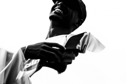 Obra de Matheus Andrés, foto em preto e branco, retrata um homem sorridente, usando boné, sob fundo branco