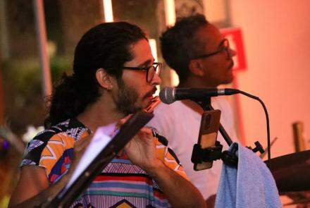 Em primeiro plano, um homem branco de óculs, cabelo comprido amarrado e baba, aparece tocando um instrumento de sopro. Ao fundo há um homem negro de óculos e camiseta branca