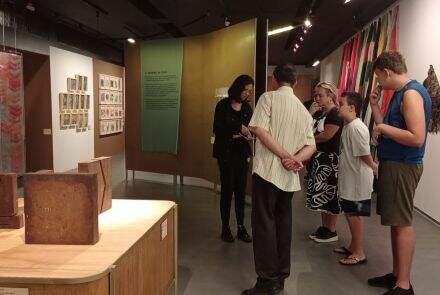 grupo de cinco pessoas, dois homens, uma criança e duas mulheres, observam um exposição em no museu. São quadros e blocos