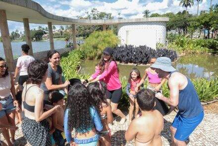 grupo de crianças e adultos brincam com baldes de água, em um dia ensolarado. Ao fundo há a estrutura e jardins da Casa do Baile