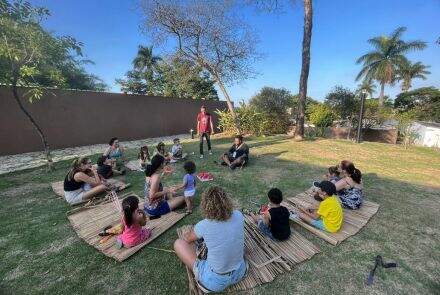 grupo de crianças e alguns adultos, todos sentados em esteiras de bambu, nos gramados do jardim MCK