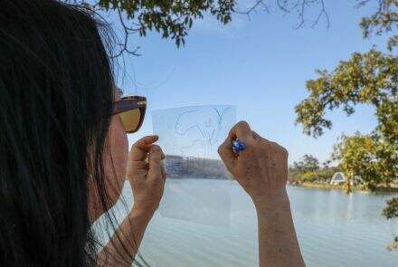 Uma mulher, de costas para câmera, segura um papel transparente e faz um desenho nele. Ela está posicionada de frente para a Lagoa da Pampulha 