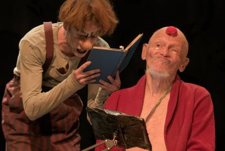 Na imagem, dois atores são retratados em uma cena que um deles está imerso na leitura, expressando concentração, enquanto o outro parece confuso, segurando o livro com uma expressão de dúvida. 