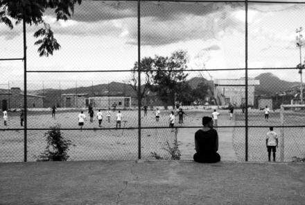 Imagem em preto e branco, com mulher sentada em primeiro plano observando um jogo de futebol em um campo de várzea