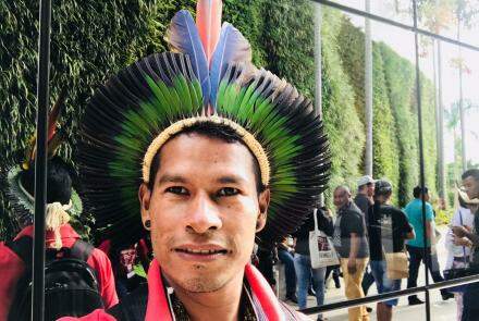 Fotografia colorida de Siwe, homem indígena com cocar na cabeça, ao fundo outras pessoas.