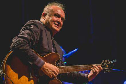 Foto colorida de Juarez Moreira em apresentação no palco. Ele está sentado, com um instrumento do tipo violão nas mãos e esboçando um sorriso.