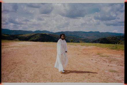 Fotografica colorida de Rico Dalasam. Ele está em pé, vestido com um túnica branco. Ao fundo, uma paisagem de serra. fotografia diurna com ceu parcialmente aberto.