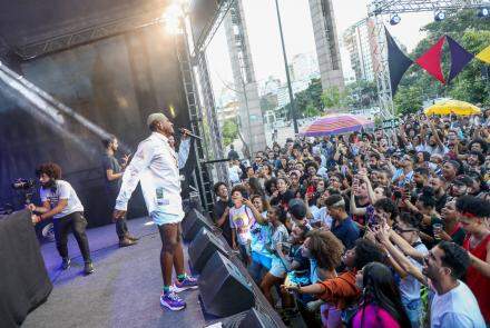 Foto de um show musical lotado. A foto mostra um palco ao ar livre, na praça da Estação, em Belo Horizonte. No palco, um homem negro, de cabelos curtos tingidos de loiro, vestindo uma blusa social branca, saia jeans branca e tênis roxo. O homem está segurando um microfone e cantando para uma plateia lotada. 
