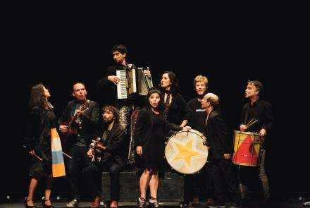 Foto do Grupo Galpão cantando e tocando em frente a um fundo preto. Seis atores e três atrizes vestidos com roupas sociais pretas cantam e tocam instrumentos, como tambor, acordeon e violão.