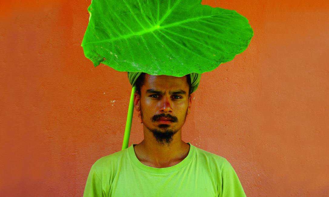 Imagem mostra o artista Paulo Nazareth com uma blusa verde limão e uma folha de taioba na cabeça. Ao fundo uma parede laranja.