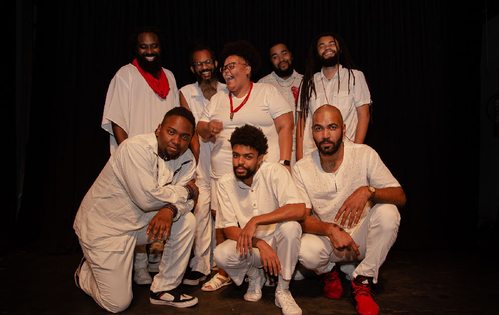 Quatro homens negros em pé vestidos de branco, uma mulher negra vestida de branco em pé e três homens negros agachados vestidos de branco