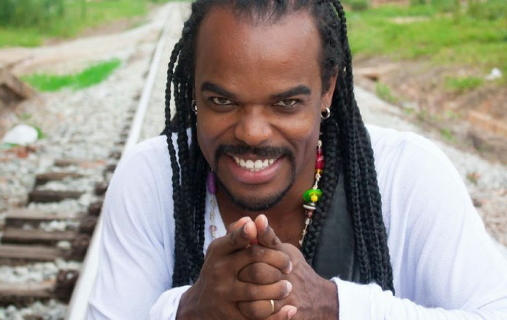 Foto: Divulgação Tom Nascimento, homem negro de camisa branca. Eles está sorrindo e com as mão unidas . Tom usa um colar
