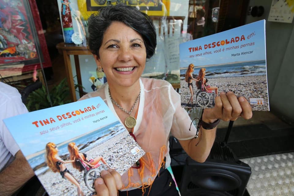 Foto: Arquivo Pessoal Marta Alencar, criadora da personagem cadeirante Tina Descolada, segurando seu livro sobre histórias da Tina