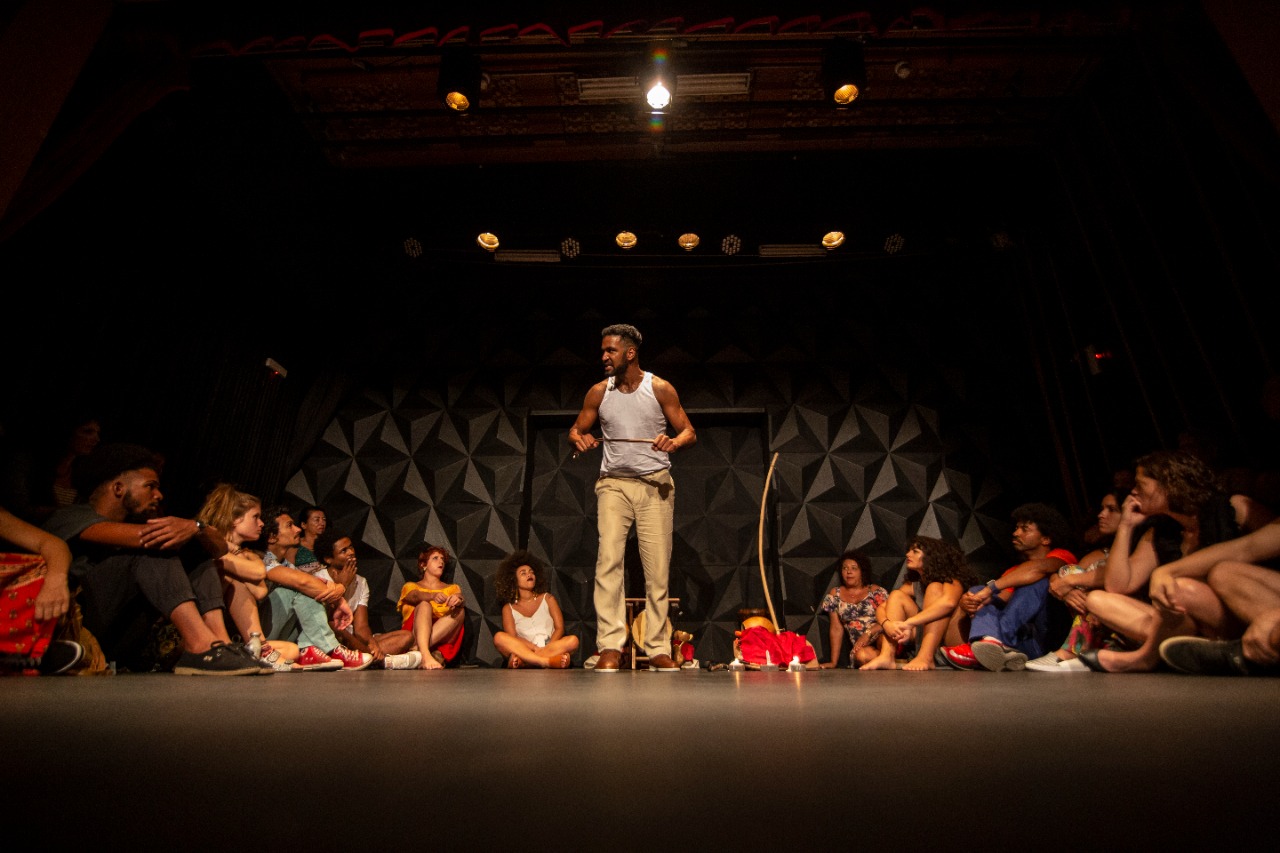 Fotografia de Rodrigo Negão no centro do palco, com pessoas sentadas ao redor durante uma apresentação do Espetáculo Mata Rasteira.