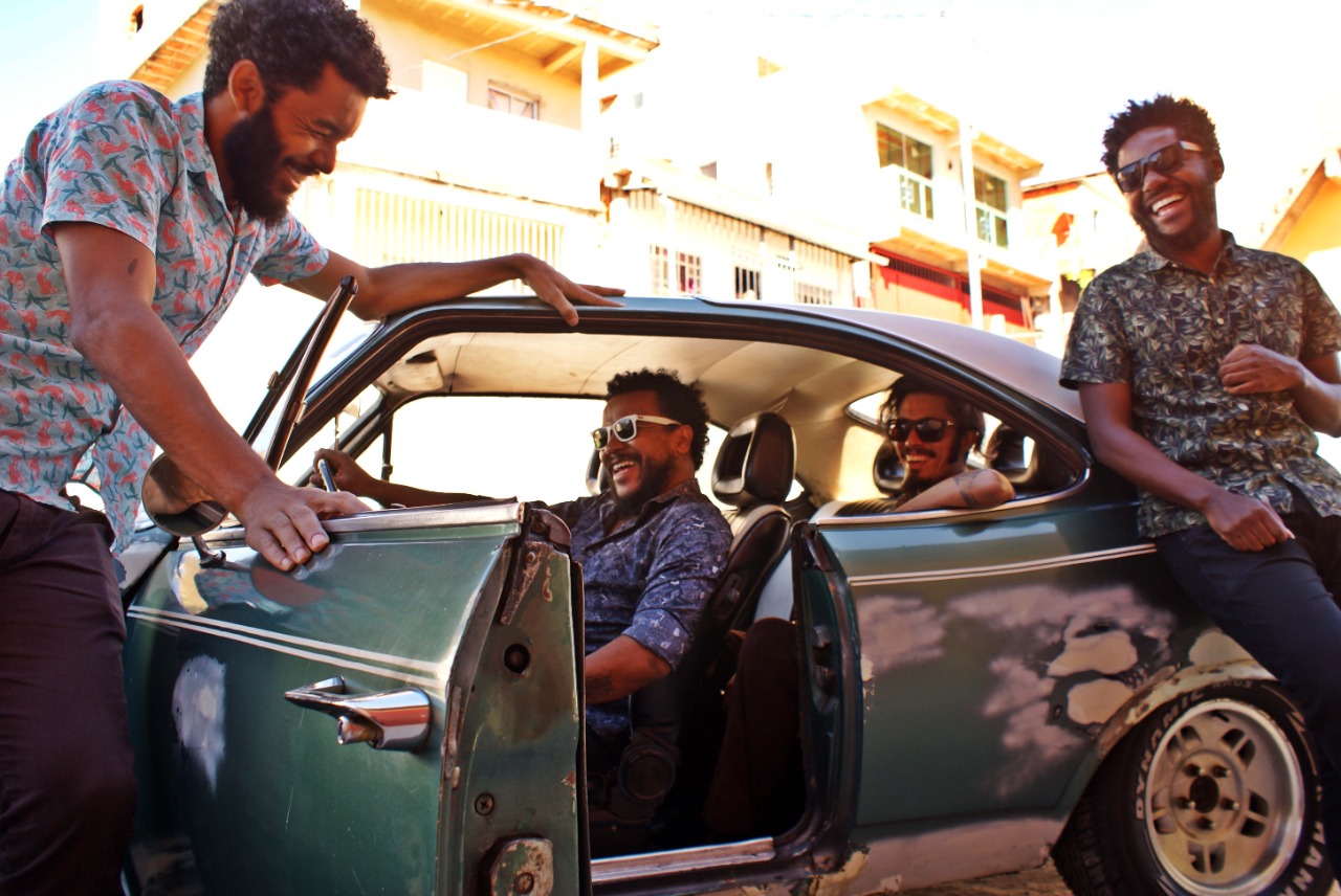 Anna Lages/Divulgação Foto com carro em primeiro plano, de portas abertas, com dois músicos dentro dele, e outros dois um de cada lado do automóvel