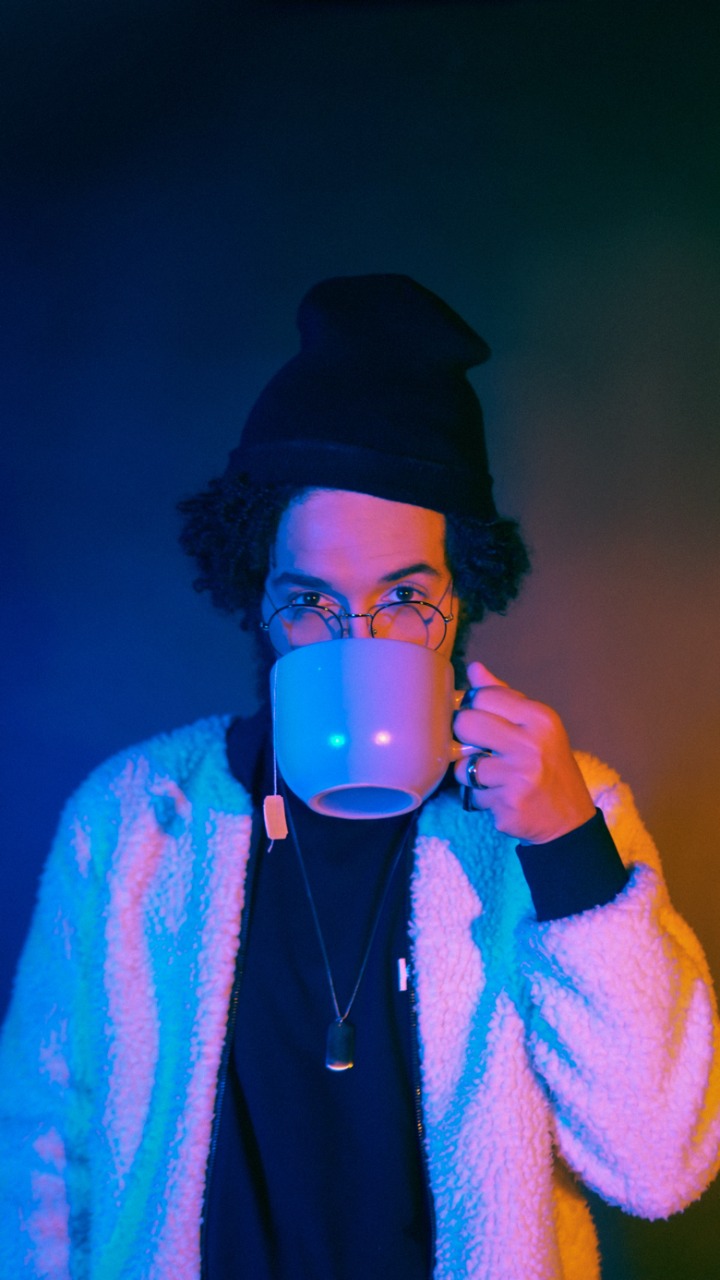 Foto de divulgação da banda belo-horizontina Dom Pepo. Na imagem um homem branco, de cabelos pretos, altos acima da orelha, veste touca de lã preta, óculos de grau, jaqueta branca com punho preto e blusa preta por dentro. O homem olha para a câmera enquanto bebe uma xícara de chá. A xícara de chá é cinza e grande, tampa sua boca e parte do seu nariz. Fim da descrição.