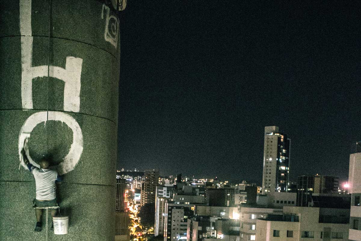 A foto mostra a cidade sob o ângulo de cima de uma prédio. Um homem está de costas e pendurado em um dos prédios, pintando uma empena. Na imagem há prédios, ruas, carros, árvores e luzes. 