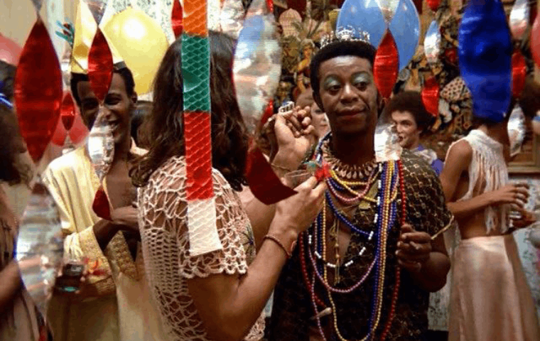Foto de uma cena do filme "A Rainha Diaba", composta por um ambiente festivo, com balões de São João pendurados. Na cena, vários atores e atrizes, com destaque para o personagem Diaba.