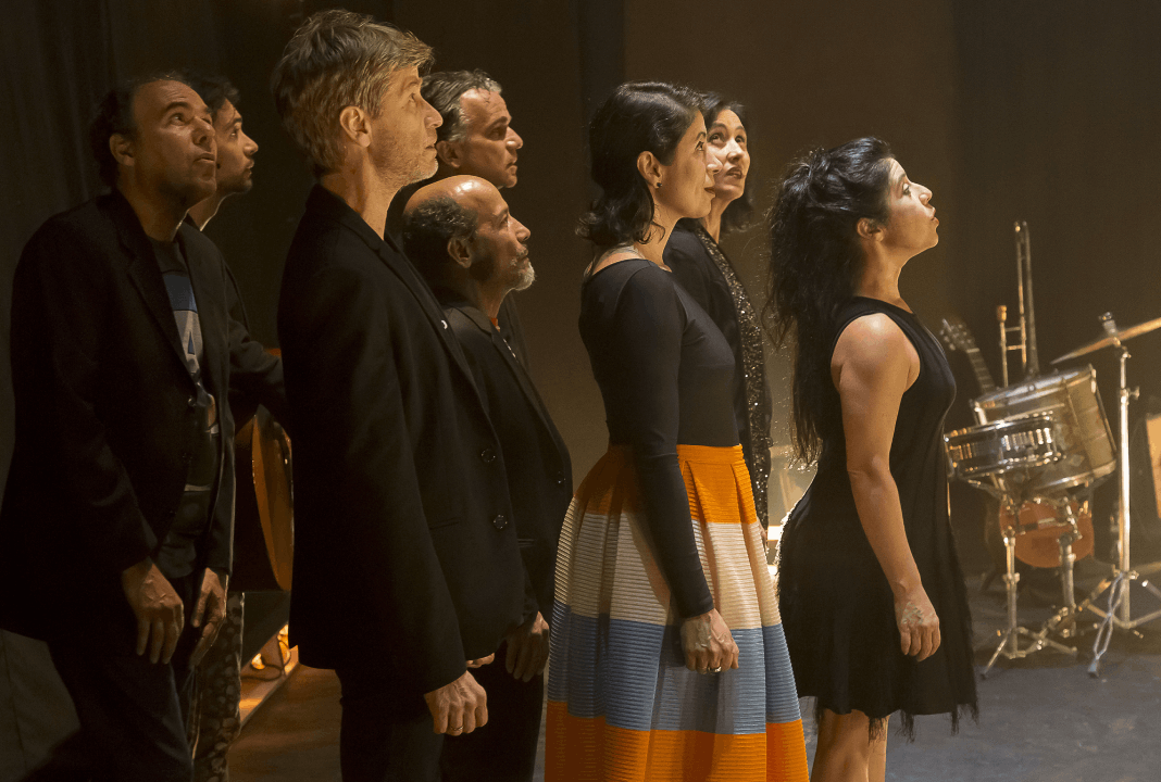 Foto registrada na lateral do palco, com os integrantes de De Tempos Somos olhando para a direção do público. 5 homens atrás e 3 mulheres à frente. Todos vestidos com roupas pretas, exceto uma mulher com uma saia com faixas amarela, azul e branco.
