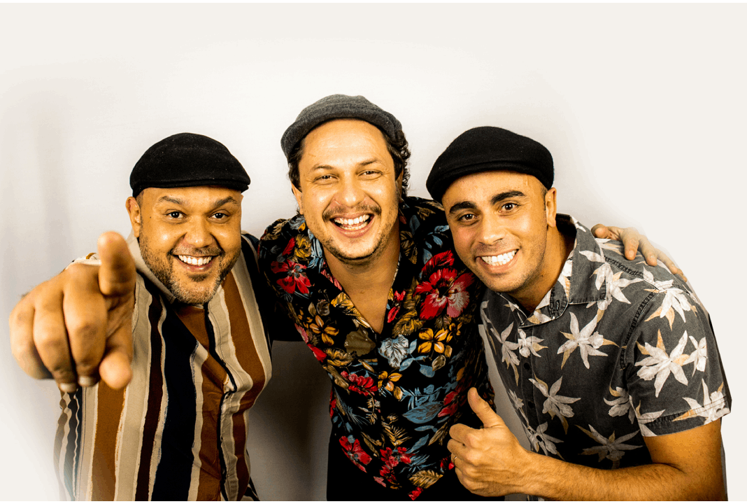 Foto em fundo branco com os integrantes do Trio Lampião, três homens se abraçando e sorridentes, olhando para a câmera.
