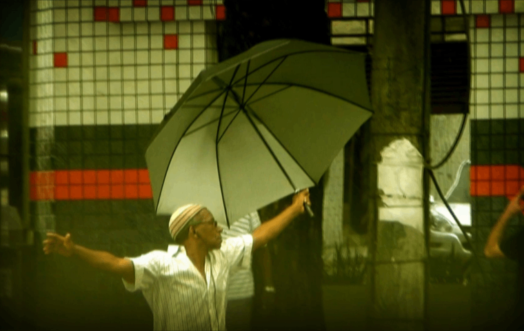 Foto: Reprodução Imagem de um senhor negro, segurando um guarda-chuvas, de braços abertos numa rua.