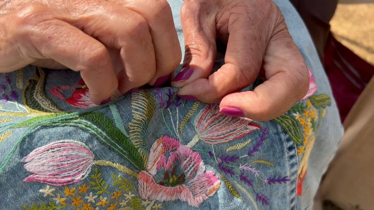 A imagem mostra duas mãos fazendo um bordado de rosas em um tecido azul 