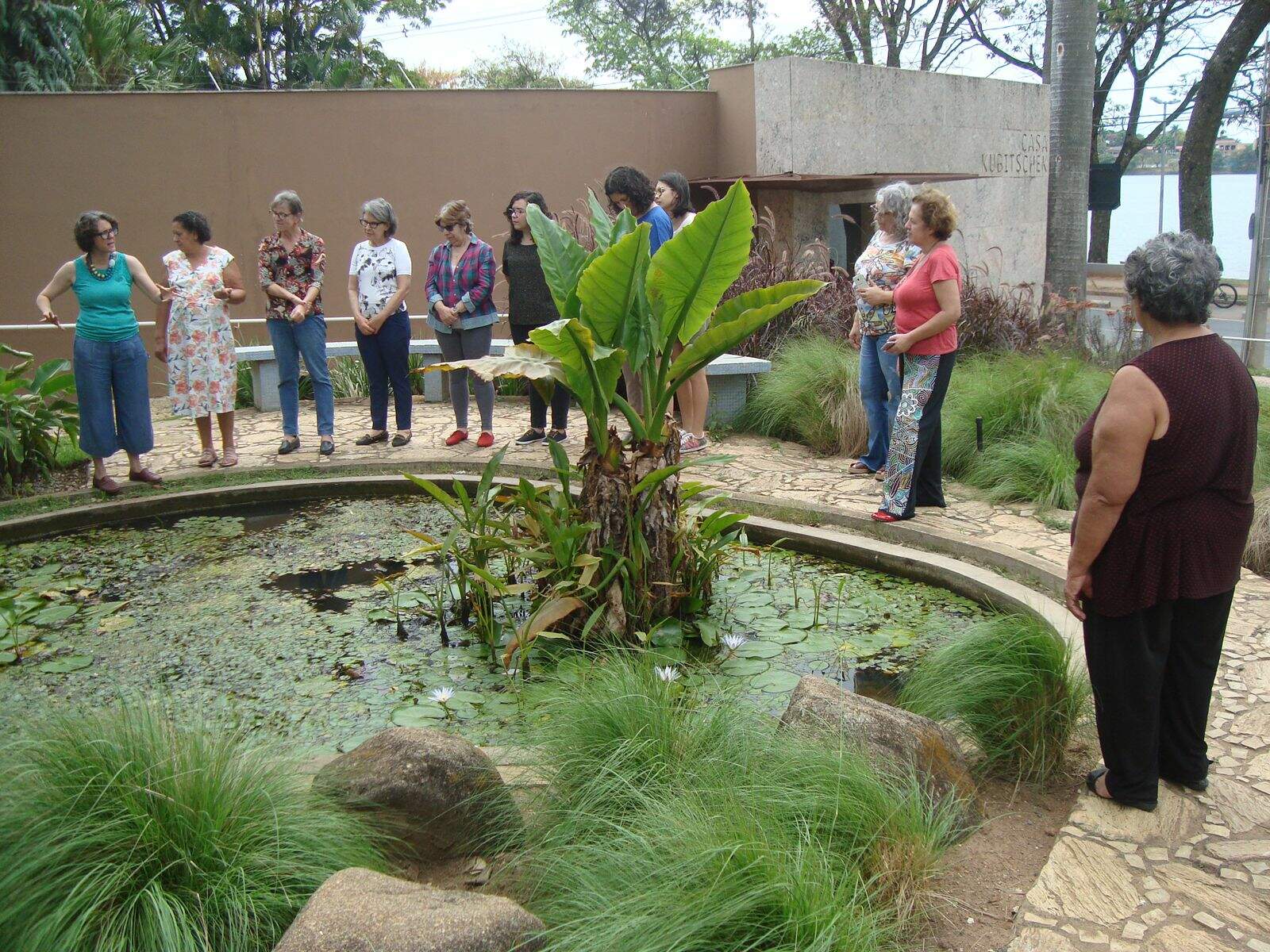 Grupo de pessoas observam um jardim. Na imagem as pessoas estão posicionadas ao redor de um pequeno lago no jardim da MCK. É possível notar plantas verdes ao redor.