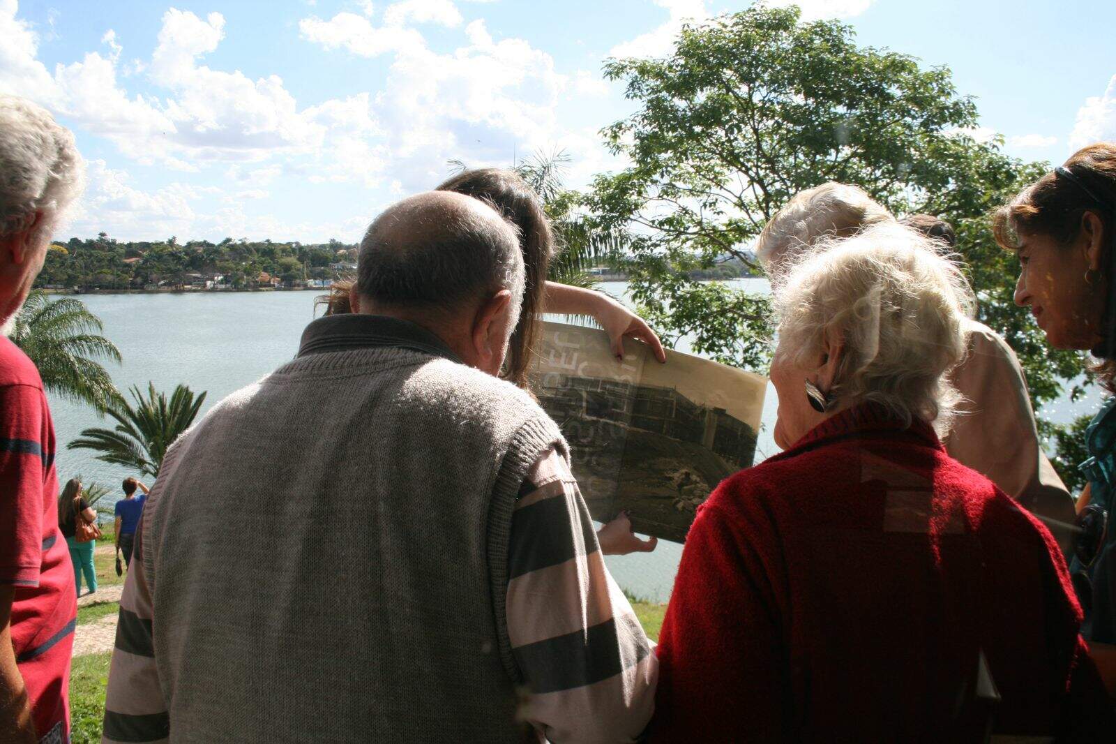 Um grupo de idosos observa uma fotografia em preto e branco. Eles estão de costas para câmera e de frente para a Lagos da Pampulha, que aparece ao fundo. 