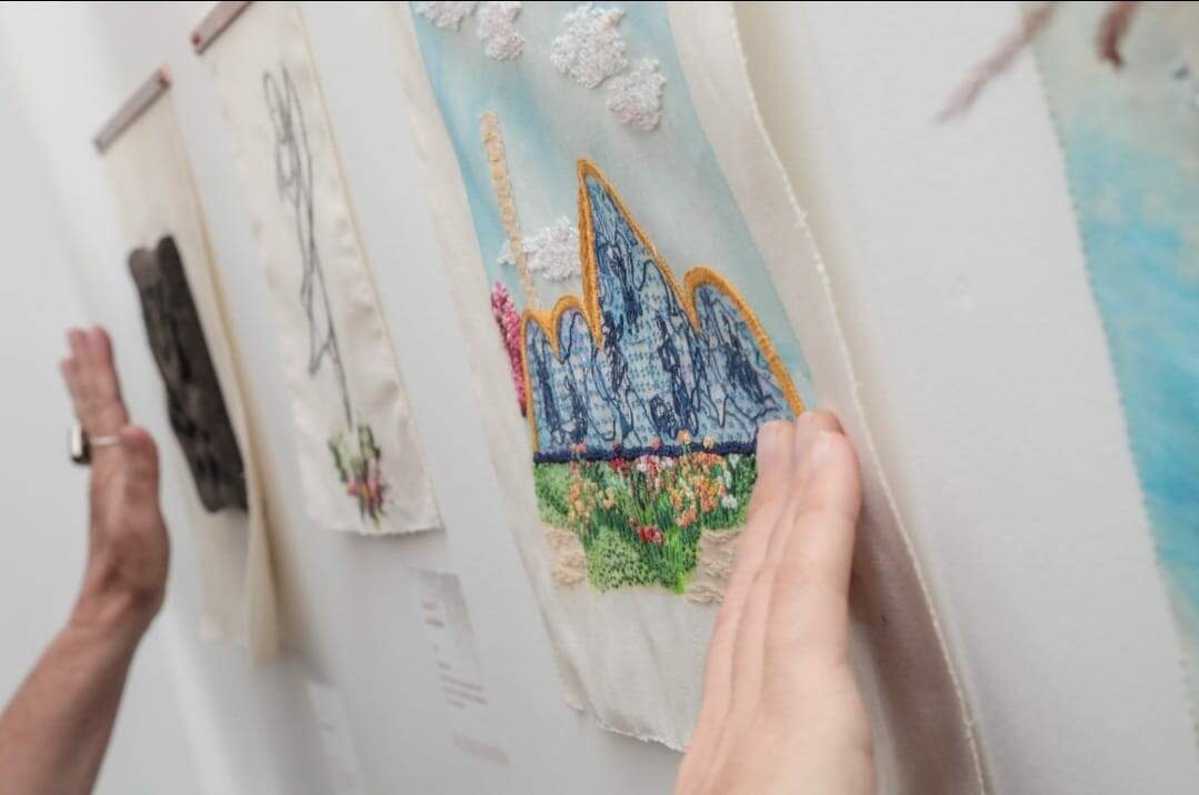 A foto revela uma mão branca encostando em uma peça de bordado pendurada na perede. A peça apresenta a imagem da igreja da Pampulha bordada em azul 