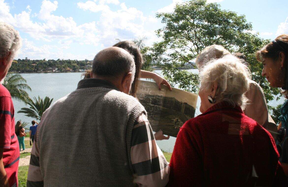 Um grupo de cinco idosos admiram um croqui apresentado por uma mulher. Eles estão de costas para câmera. No centro da imagem está um senhor de pulôver cinza e uma senhora de camisa vermelha. 