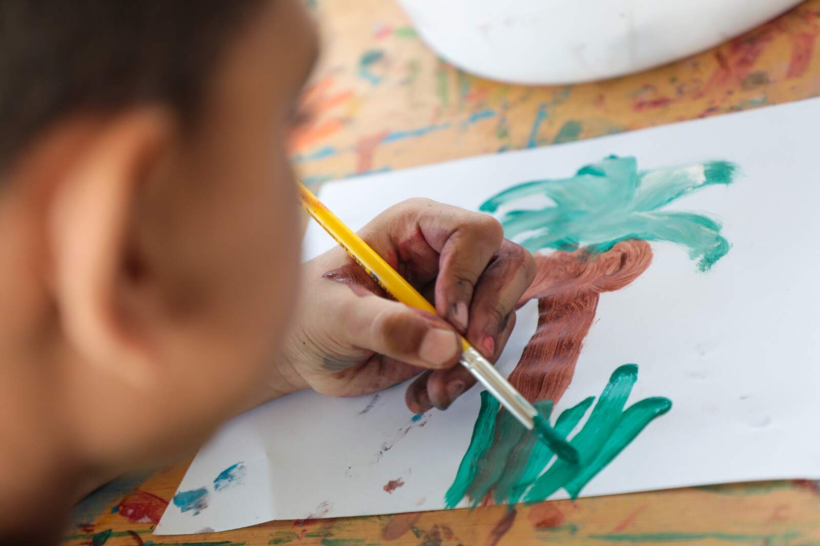 uma criança, que aparece desfocada na imagem, pinta um papel em branco. O desenho revela um coqueiro.
