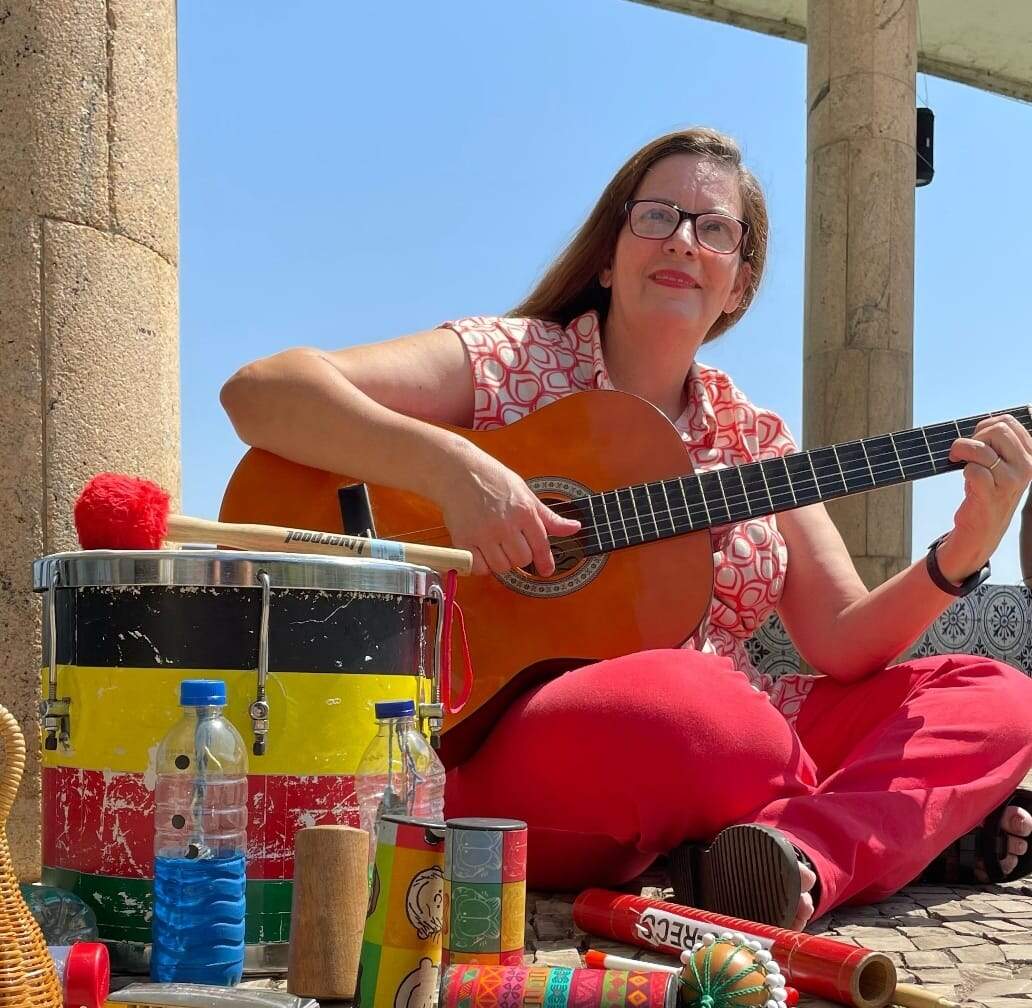 Alguns instrumentos musicais coloridos estão apoiados no chã, como um tambor. Uma mulher de roupa rosa está senta um pouco mais ao fundo, ela segura um violão e olha para câmera. Ela é branca, possui cabelos vermelhos e usa óculos.