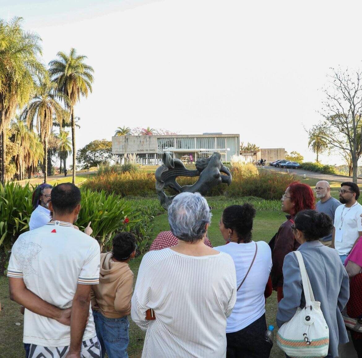Grupo de pessoas de costas para a câmera observam ao jardim e fachada do Museu de Arte da Pampulha, localizado ao fundo da imagem.