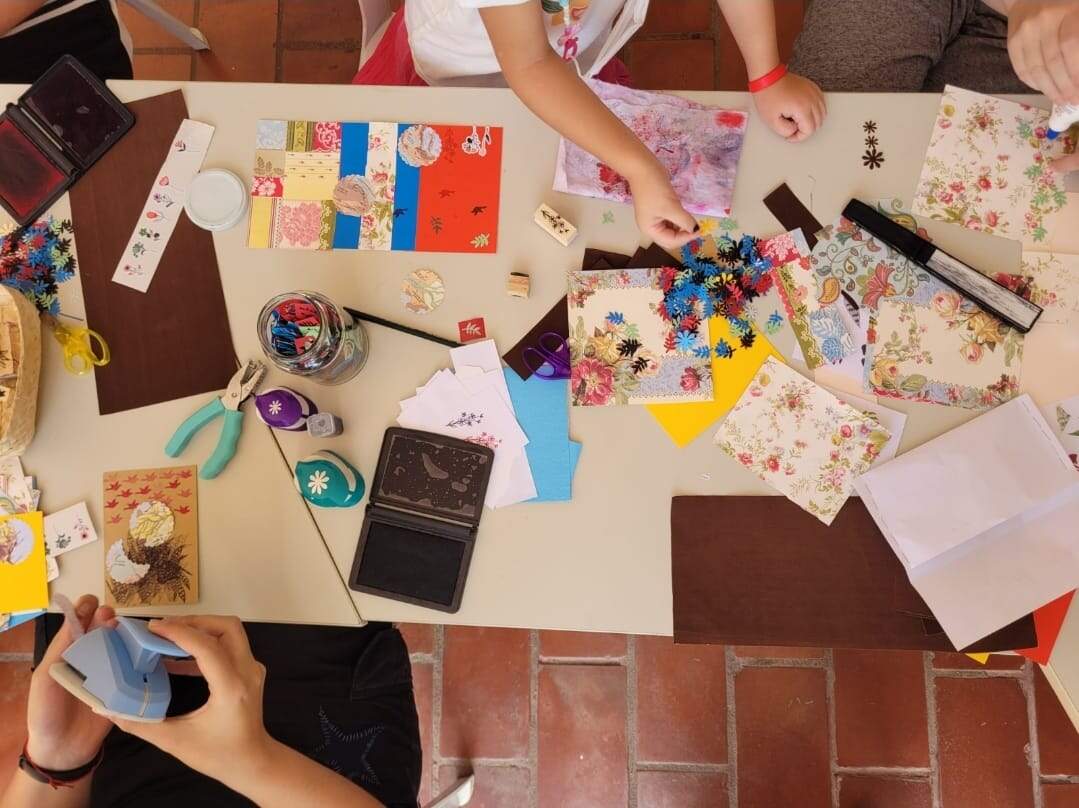 Mesa com diversos recortes coloridos e ferramentas para artesanato, como alicate, régua, tesoura, cola. Mãos de duas pessoas brancas aparecem manuseando os materiais. 