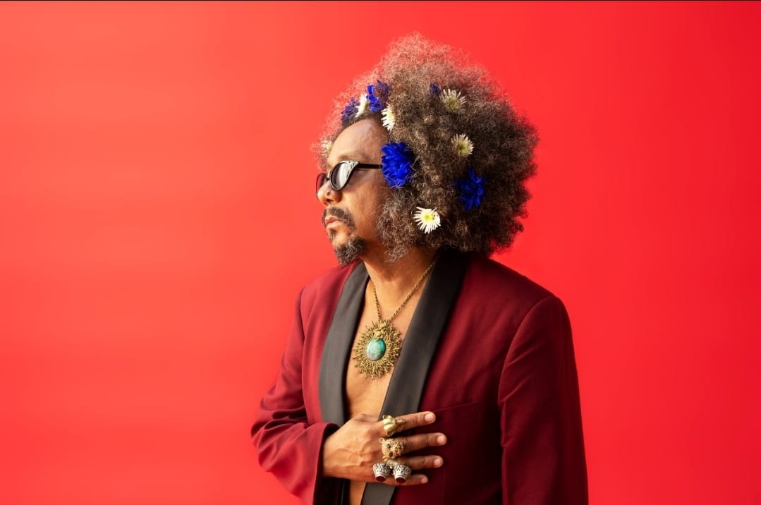 Em um fundo alaranjado, o cantor Chico César está com o corpo virado para a parte esquerda da foto. Chico é um homem negro, usando um blazer vinho, sem camisa por baixo. Ele usa um colar dourado com uma pedra azul no meio e óculos escuro. Ele está com os cabelos soltos, um black power decorado com flores azuis e brancas.