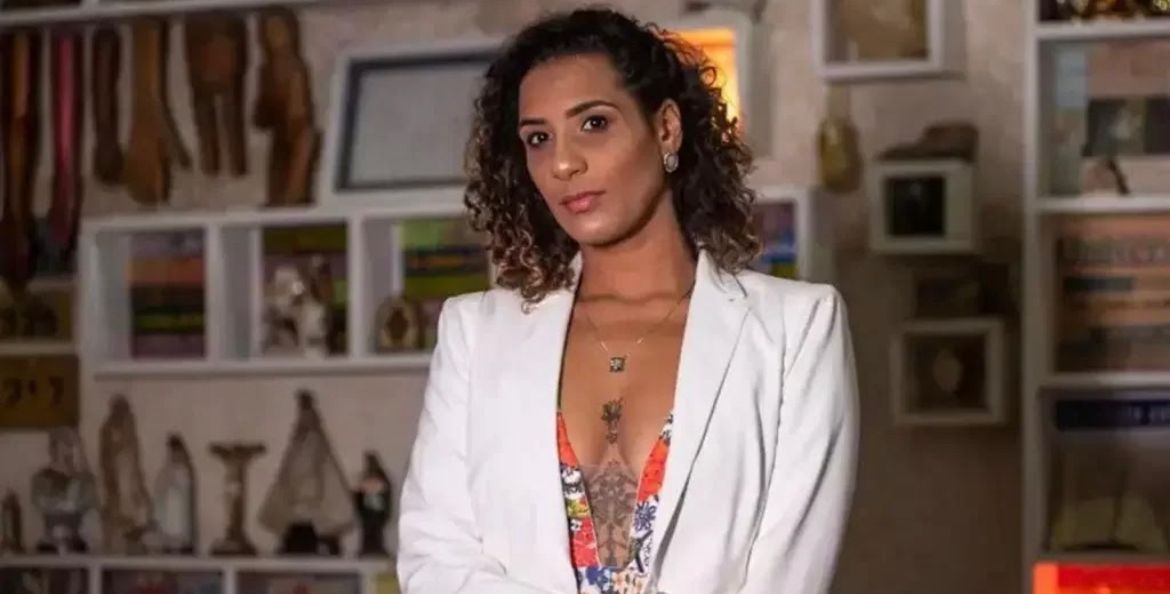 Mulher negra de blazer branco. Ela está com os cabelos soltos e olhando para câmera. 