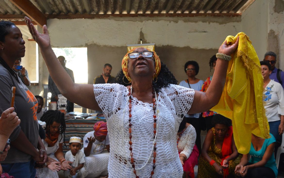 mulher negra com roupa de renda branca, colares e faixa amarela na cabeça, ergue os braços e olha para o céu