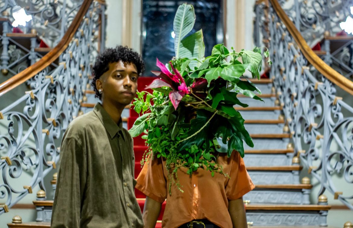 Uma pessoa negra, com camisa verde e cabelos curtos olha para câmera. Ela está em frente a uma escada. Um pouco mais ao fundo, na lateral direita, uma pessoa de camisa laranja está com algum ornamento de plantas e flores tampando o rosto