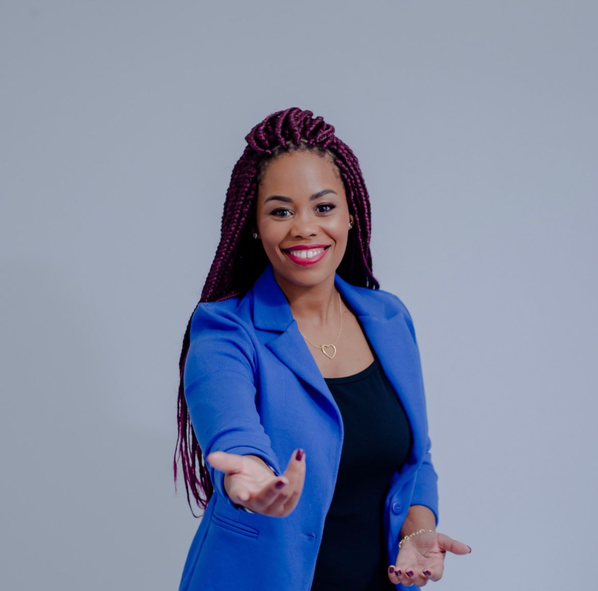 Mulher negra com cabelos de tranças, sorri e ergue os braços para frente. Ela usa calça e camisa preta, além de um blazer azul