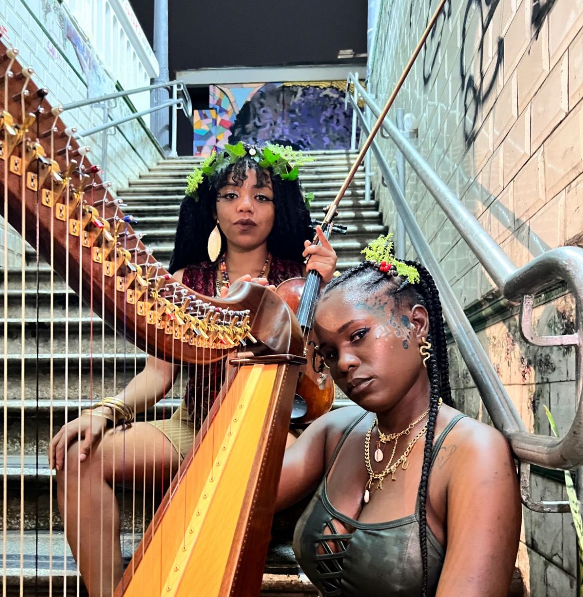 Duas mulheres negras, segurando uma harpa, olham para câmera enquanto estão sentadas em uma escada. Elas usam algumas plantas no cabelo, como adereço 